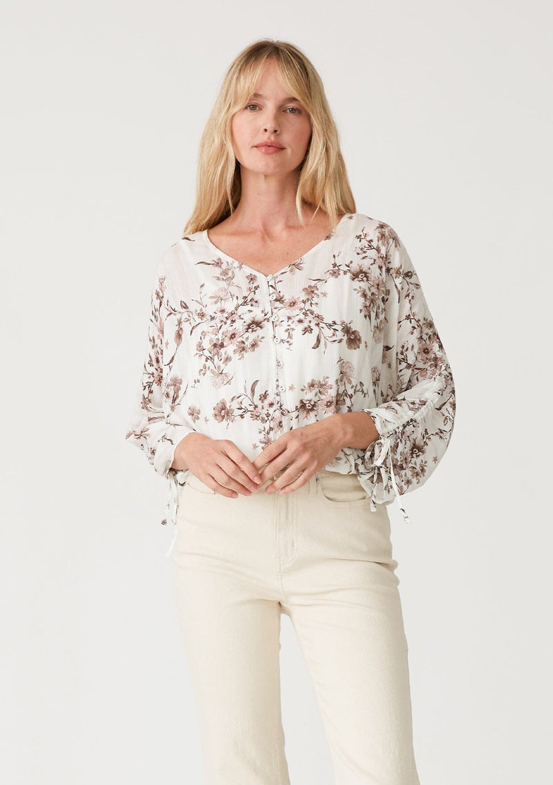 [Color: Ivory/Taupe] A front facing image of a blonde model wearing a lightweight spring top in a white and taupe purple floral print and metallic thread detail. With three quarter length sleeve, a gathered sleeve detail with ties, a v neckline, and a self covered button front. 