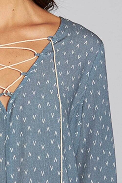 [Color: Slate Blue/Vanilla] A detail image of a brunette model wearing a blue geometric print blouse with a lace up front, tassel detail, and flowy bell sleeves. 