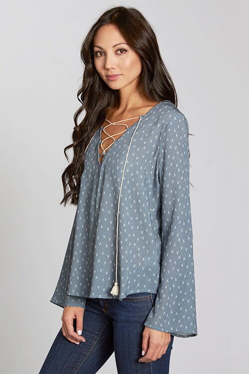 [Color: Slate Blue/Vanilla] A side facing image of a brunette model wearing a blue geometric print blouse with a lace up front, tassel detail, and flowy bell sleeves. 