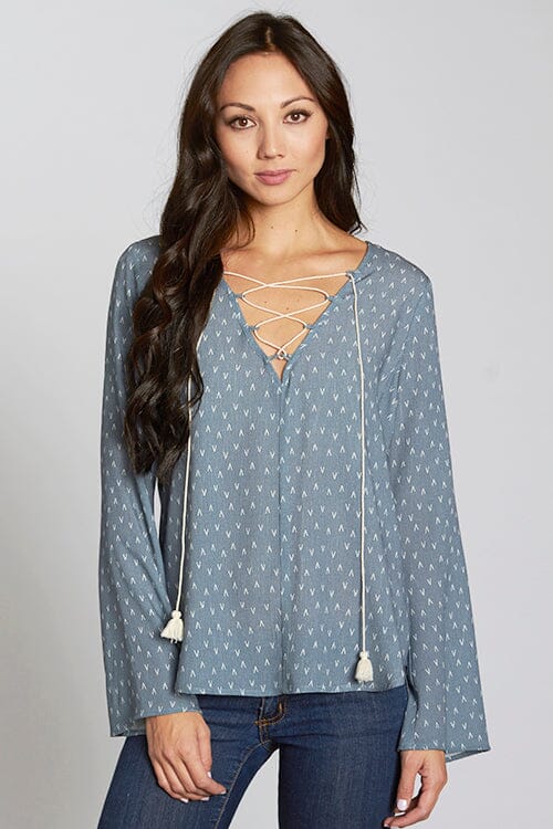 [Color: Slate Blue/Vanilla] A front facing image of a brunette model wearing a blue geometric print blouse with a lace up front, tassel detail, and flowy bell sleeves. 