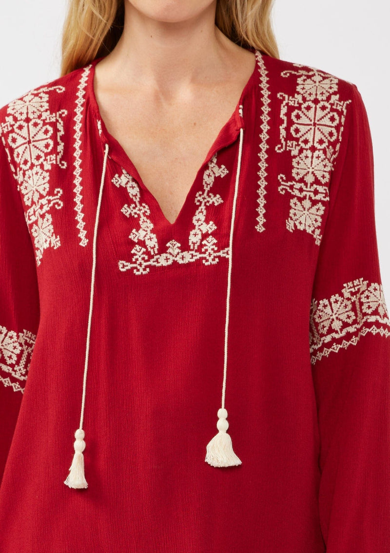[Color: Red] A detail image of a blonde model wearing a red embroidered boho peasant blouse with a split v-neckline, long sleeves, and tassel ties. 
