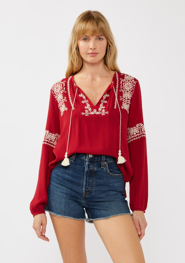[Color: Red] A front facing image of a blonde model wearing a red embroidered boho peasant blouse with a split v-neckline, long sleeves, and tassel ties. 