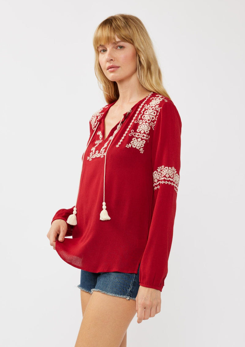 [Color: Red] A side facing image of a blonde model wearing a red embroidered boho peasant blouse with a split v-neckline, long sleeves, and tassel ties. 