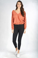 [Color: Terracotta] A front facing image of a brunette model wearing a orange boho blouse with swiss dots and embroidered eyelet sleeves. Finished with a chic tie back detail and a comfortable elastic waistline. 