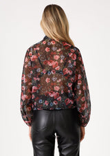 [Color: Black/Rose] A back facing image of a blonde model wearing a sheer chiffon tie front top in a black and pink floral print. With voluminous long dolman sleeves, a plunging v neckline, and a tie front waist detail that can be styled in multiple ways. 