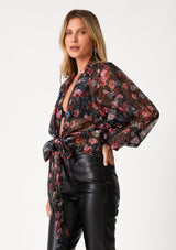 [Color: Black/Rose] A side facing image of a blonde model wearing a sheer chiffon tie front top in a black and pink floral print. With voluminous long dolman sleeves, a plunging v neckline, and a tie front waist detail that can be styled in multiple ways. 