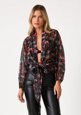 [Color: Black/Rose] A front facing image of a blonde model wearing a sheer chiffon tie front top in a black and pink floral print. With voluminous long dolman sleeves, a plunging v neckline, and a tie front waist detail that can be styled in multiple ways. 