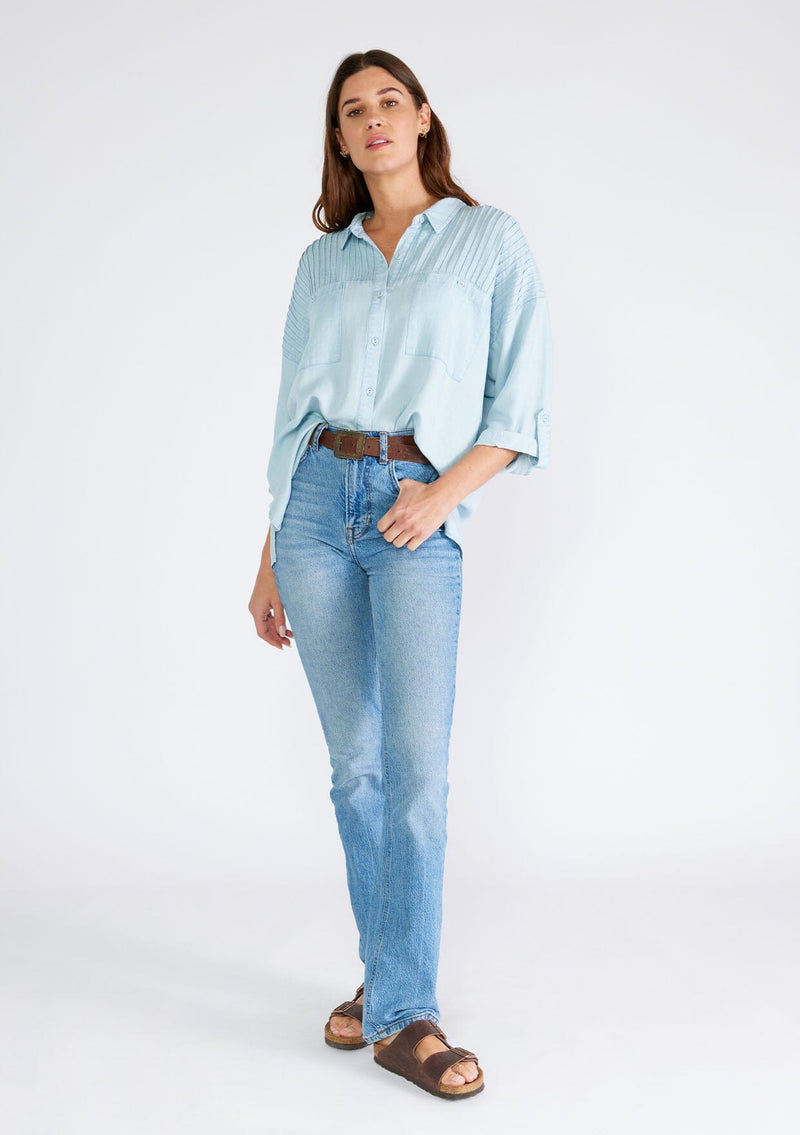 [Color: Bleach Wash] A full body front facing image of a brunette model wearing a light blue wash Tencel shirt. With long three quarter length sleeves, a button tab sleeve, a collared neckline, a button front, two front patch pockets, and pintuck details. 