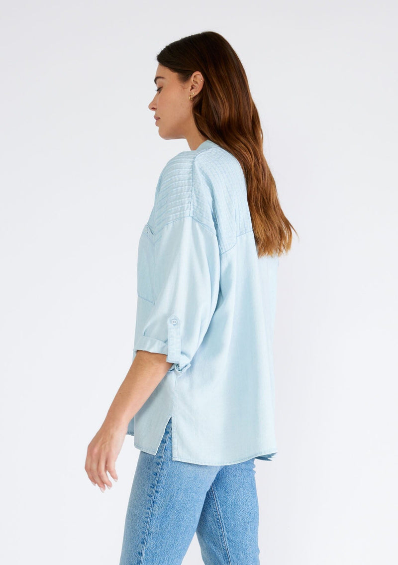 [Color: Bleach Wash] A side facing image of a brunette model wearing a light blue wash Tencel shirt. With long three quarter length sleeves, a button tab sleeve, a collared neckline, a button front, two front patch pockets, and pintuck details. 