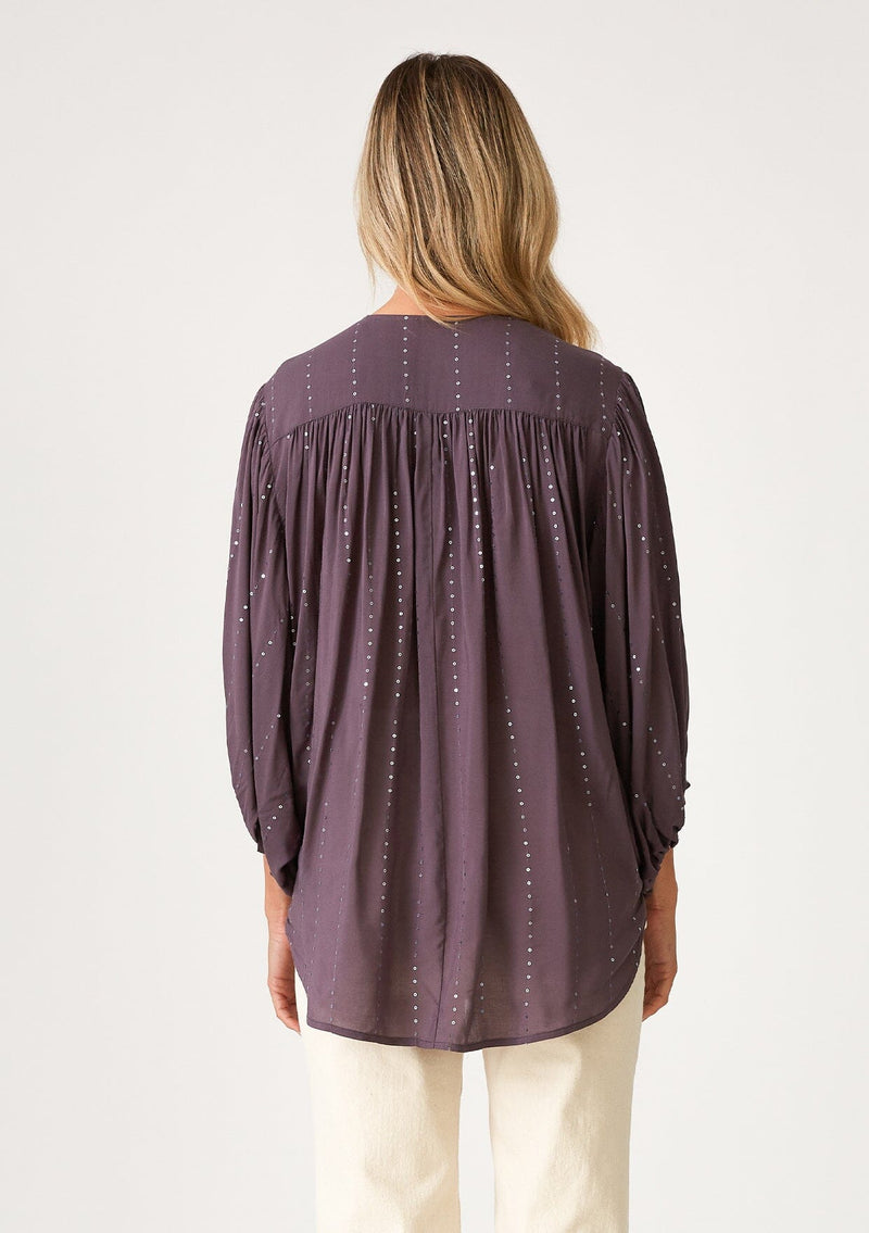 [Color: Dusty Plum] A back facing image of a blonde model wearing a dusty purple bohemian blouse with a sparkly sequin stripe. With voluminous long three quarter length sleeves, a surplice v neckline, a high low hemline, and an ultra flowy fit. 