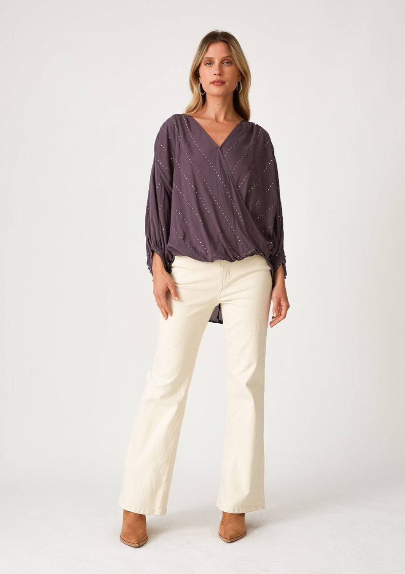 [Color: Dusty Plum] A full body front facing image of a blonde model wearing a dusty purple bohemian blouse with a sparkly sequin stripe. With voluminous long three quarter length sleeves, a surplice v neckline, a high low hemline, and an ultra flowy fit. 