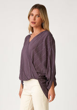 [Color: Dusty Plum] A side facing image of a blonde model wearing a dusty purple bohemian blouse with a sparkly sequin stripe. With voluminous long three quarter length sleeves, a surplice v neckline, a high low hemline, and an ultra flowy fit. 