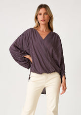 [Color: Dusty Plum] A front facing image of a blonde model wearing a dusty purple bohemian blouse with a sparkly sequin stripe. With voluminous long three quarter length sleeves, a surplice v neckline, a high low hemline, and an ultra flowy fit. 