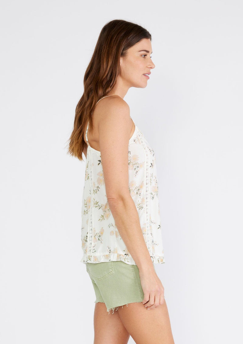[Color: Ivory/Dusty Coral] A side facing image of a brunette model wearing a flowy bohemian camisole in an ivory and pink floral print. With adjustable spaghetti straps, a v neckline, lace trim, a ruffled hemline, and pintuck details. 