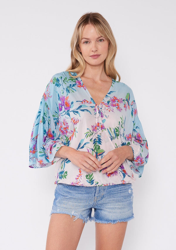 [Color: Light Pink/Purple] A blonde model wearing a vibrant tropical floral print top. This summer top features an ombre effect, surplice v neckline, front tie closure, front elastic hem, and long flowy wide sleeves. A vacation top paired with denim shorts. 