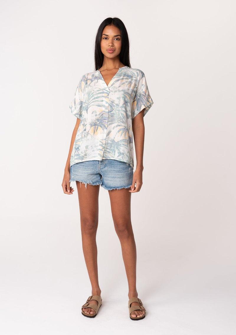 [Color: Natural/Seafoam] A full body front facing image of a brunette model wearing a lightweight summer tee in a green palm leaf print. With short cuffed sleeves, a v neckline, and a relaxed fit. 