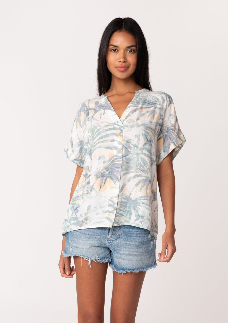 [Color: Natural/Seafoam] A front facing image of a brunette model wearing a lightweight summer tee in a green palm leaf print. With short cuffed sleeves, a v neckline, and a relaxed fit. 