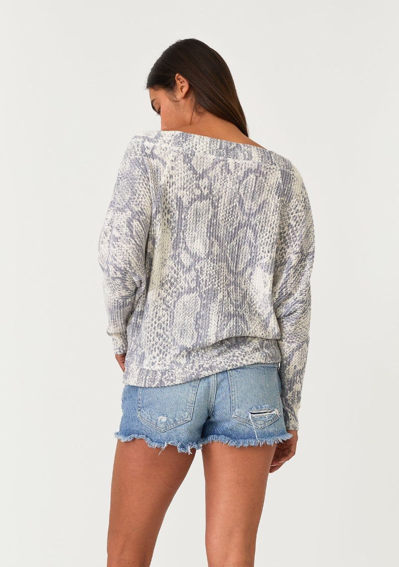 [Color: Light Grey/Ivory] A back facing image of a brunette model wearing a soft waffle knit pullover top in a grey and ivory snakeskin print. With long tapered dolman sleeves and a wide neckline that can be worn on or off the shoulder.