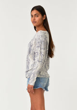 [Color: Light Grey/Ivory] A side facing image of a brunette model wearing a soft waffle knit pullover top in a grey and ivory snakeskin print. With long tapered dolman sleeves and a wide neckline that can be worn on or off the shoulder.