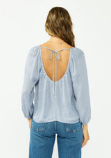 [Color: Dusty Blue] A back facing image of a brunette model wearing a blue satin jacquard blouse with a wide v neckline and a tie back detail. 
