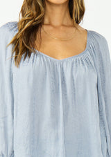 [Color: Dusty Blue] A detail image of a brunette model wearing a blue satin jacquard blouse with a wide v neckline and a tie back detail. 