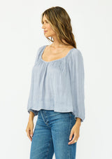[Color: Dusty Blue] A side facing image of a brunette model wearing a blue satin jacquard blouse with a wide v neckline and a tie back detail. 