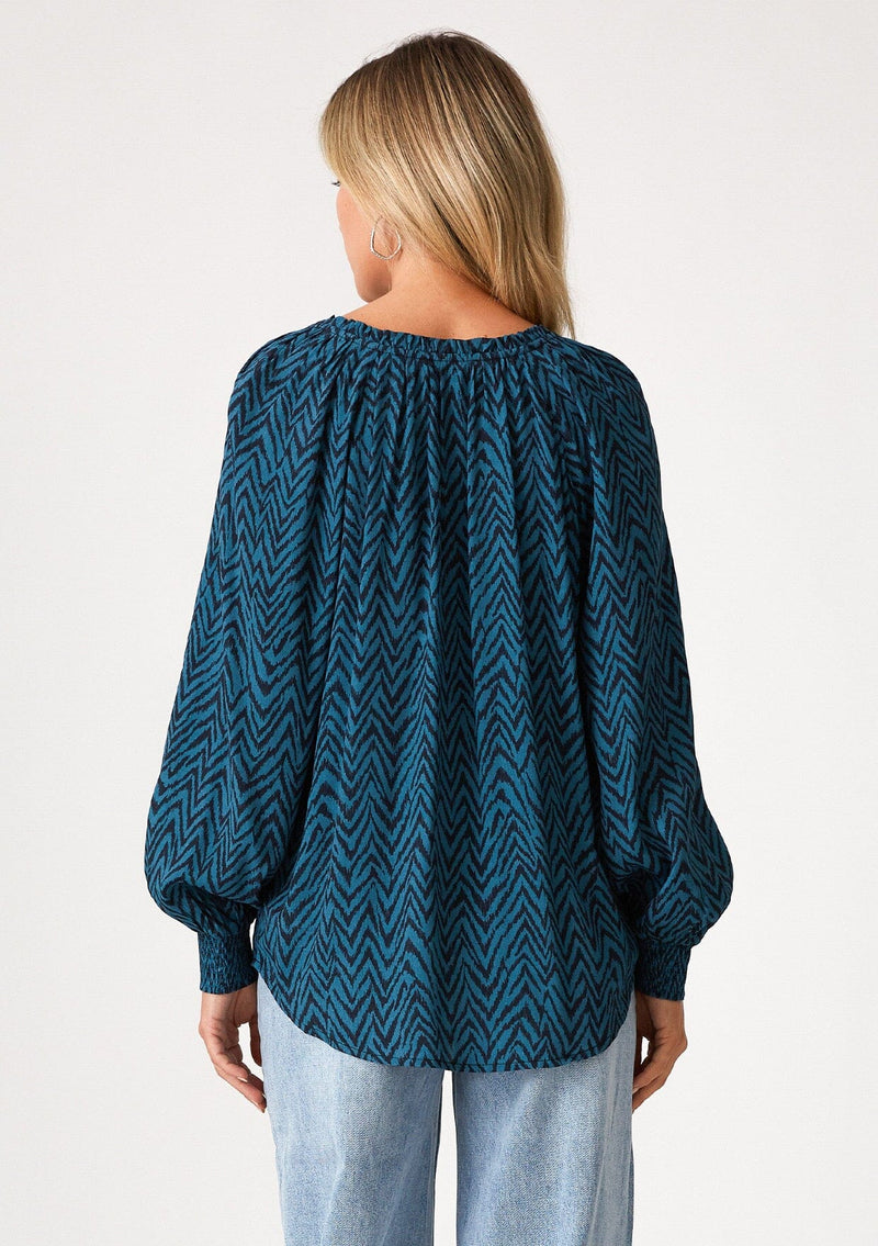 [Color: Teal/Navy] A back facing image of a blonde model wearing a bohemian fall blouse in a teal and navy blue chevron striped print. With voluminous long raglan sleeves, smocked elastic wrist cuffs, a split v neckline with tassel ties, and a flowy relaxed fit. 