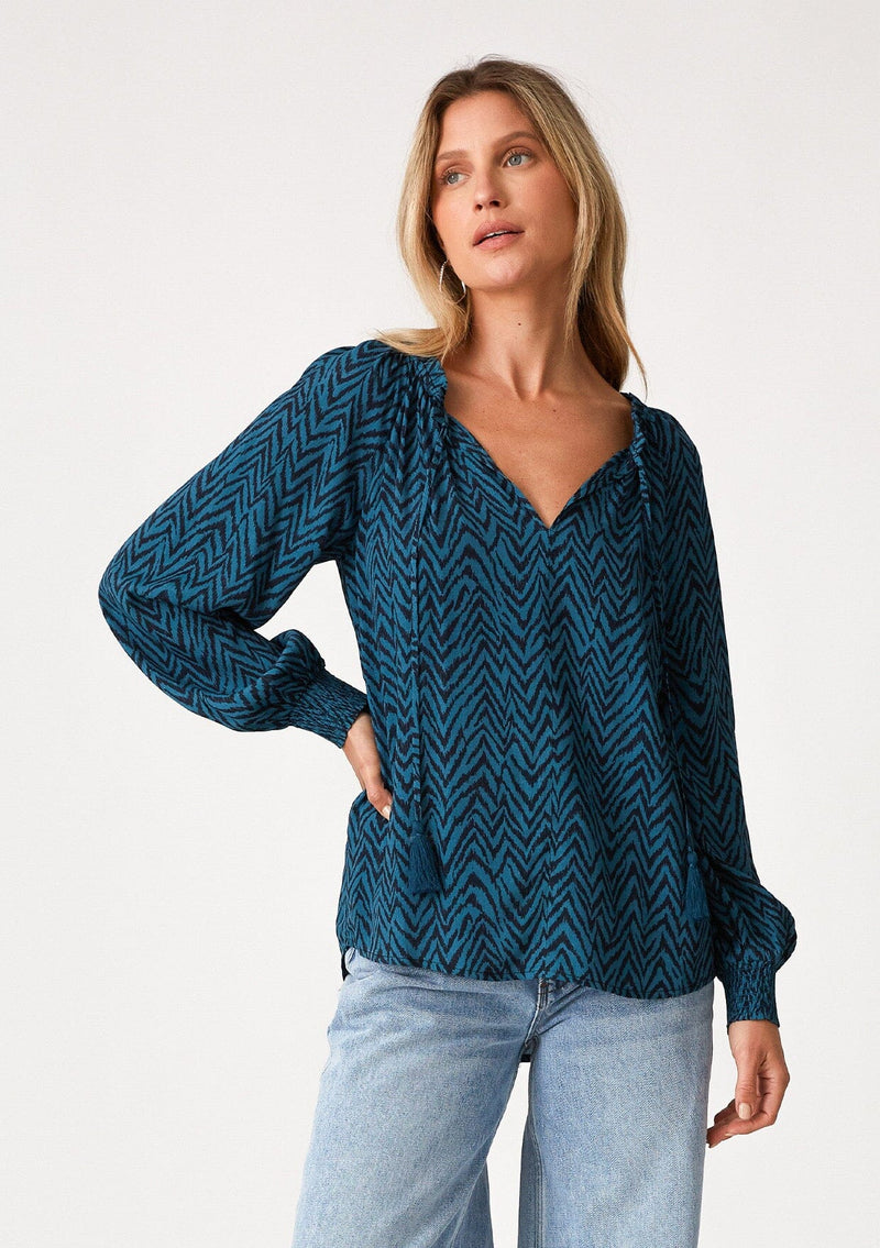 [Color: Teal/Navy] A front facing image of a blonde model wearing a bohemian fall blouse in a teal and navy blue chevron striped print. With voluminous long raglan sleeves, smocked elastic wrist cuffs, a split v neckline with tassel ties, and a flowy relaxed fit. 