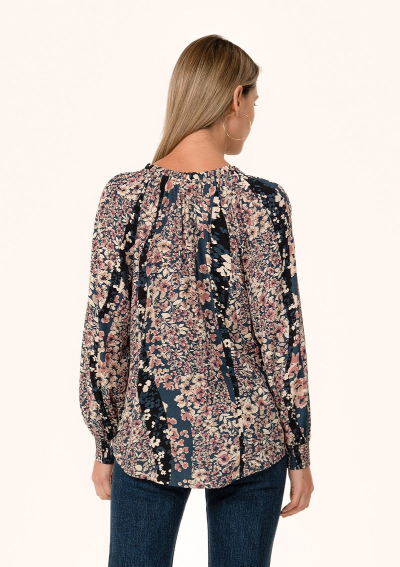 [Color: Dusty Rose/Navy] A back facing image of a blonde model wearing a bohemian fall blouse in a mixed blue and pink floral print. With voluminous long sleeves, smocked elastic wrist cuffs, a split v neckline with ties, and a relaxed fit. 