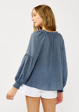 [Color: Blue] A blonde model wearing a classic lightweight sheer blue peasant top. With a ruffled neckline, tassel ties, long voluminous bishop sleeves, and a relaxed flowy fit.