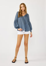 [Color: Blue] A blonde model wearing a classic lightweight sheer blue peasant top. With a ruffled neckline, tassel ties, long voluminous bishop sleeves, and a relaxed flowy fit.