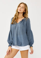 [Color: Blue] A blonde model wearing a classic lightweight sheer blue peasant top. With a ruffled neckline, tassel ties, long voluminous bishop sleeves, and a relaxed flowy fit.