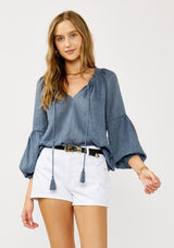 [Color: Blue] A blonde model wearing a classic lightweight sheer blue peasant top. With a ruffled neckline, tassel ties, long voluminous bishop sleeves, and a relaxed flowy fit.