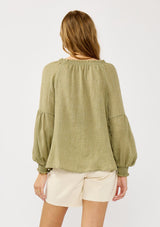 [Color: Olive] A blonde model wearing a classic lightweight sheer green peasant top. With a ruffled neckline, tassel ties, long voluminous bishop sleeves, and a relaxed flowy fit.