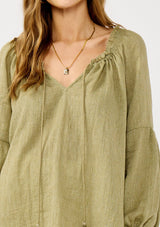 [Color: Olive] A blonde model wearing a classic lightweight sheer green peasant top. With a ruffled neckline, tassel ties, long voluminous bishop sleeves, and a relaxed flowy fit.
