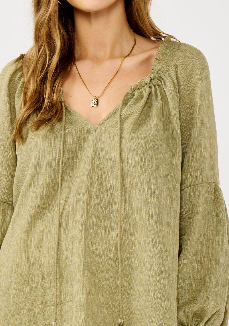 [Color: Olive] A blonde model wearing a classic lightweight sheer green peasant top. With a ruffled neckline, tassel ties, long voluminous bishop sleeves, and a relaxed flowy fit.