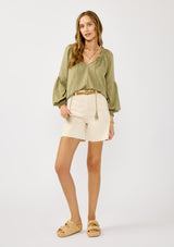 [Color: Olive] A blonde model wearing a classic lightweight sheer green peasant top. With a ruffled neckline, tassel ties, long voluminous bishop sleeves, and a relaxed flowy fit.