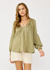 [Color: Olive] A blonde model wearing a classic lightweight sheer green peasant top. With a ruffled neckline, tassel ties, long voluminous bishop sleeves, and a relaxed flowy fit.