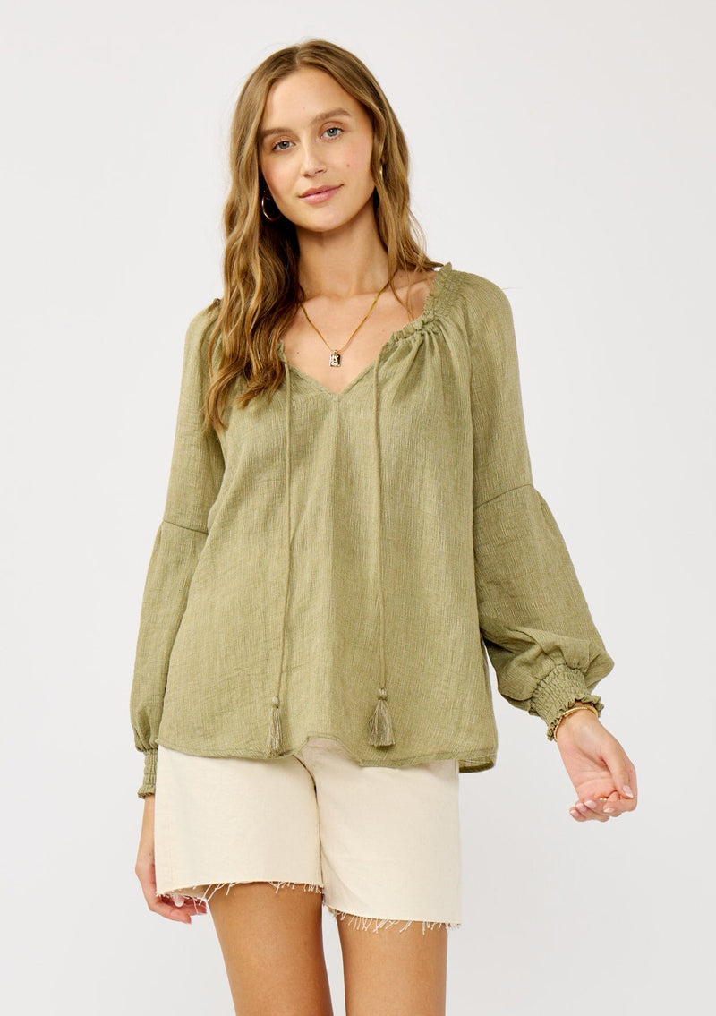 [Color: Olive] A blonde model wearing a classic lightweight sheer green peasant top. With a ruffled neckline, tassel ties, long voluminous bishop sleeves, and a relaxed flowy fit.