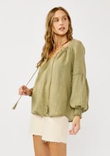 [Color: Olive] A blonde model wearing a classic lightweight sheer green peasant top. With a ruffled neckline, tassel ties, long voluminous bishop sleeves, and a relaxed flowy fit.