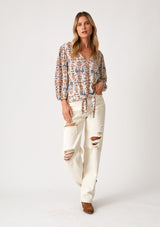 [Color: Off White/Rust] A full body front facing image of a blonde model wearing a lightweight and relaxed fall top in an off white, blue, and rust red bohemian print. With three quarter length long sleeves, a v neckline, and a tie waist detail. 