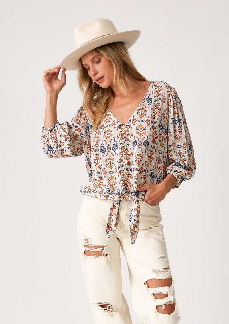 [Color: Off White/Rust] A front facing image of a blonde model wearing a lightweight and relaxed fall top in an off white, blue, and rust red bohemian print. With three quarter length long sleeves, a v neckline, and a tie waist detail. 