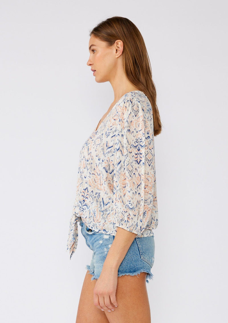 [Color: Cream/Dusty Blue] A side facing image of a brunette model wearing a lightweight bohemian blouse designed in an off white and blue print. With a v neckline, a tie waist detail, and three quarter length sleeves. 
