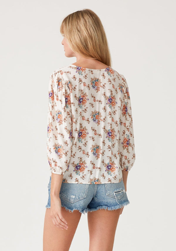 [Color: Natural/Rust] A back facing image of a blonde model wearing a best selling tie front blouse in a white and pink floral print. With three quarter length sleeves, a v neckline, and a relaxed fit. 
