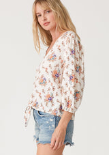 [Color: Natural/Rust] A side facing image of a blonde model wearing a best selling tie front blouse in a white and pink floral print. With three quarter length sleeves, a v neckline, and a relaxed fit. 