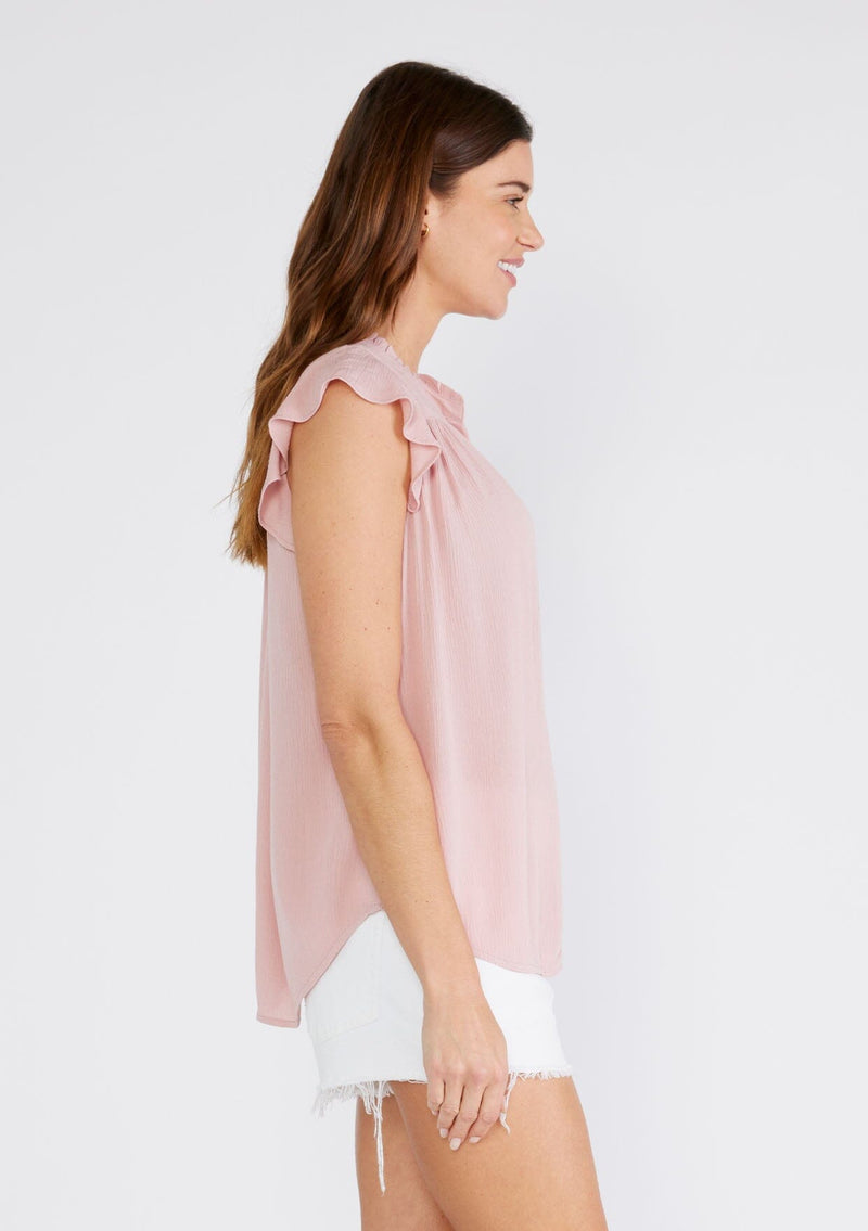 [Color: Dusty Pink] A side facing image of a brunette model wearing a soft pink crinkle gauze short sleeve flutter top with a button front and a ruffled neckline.