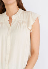 [Color: Natural] A close up front facing image of a brunette model wearing an off white crinkle gauze short sleeve flutter top with a button front and a ruffled neckline.