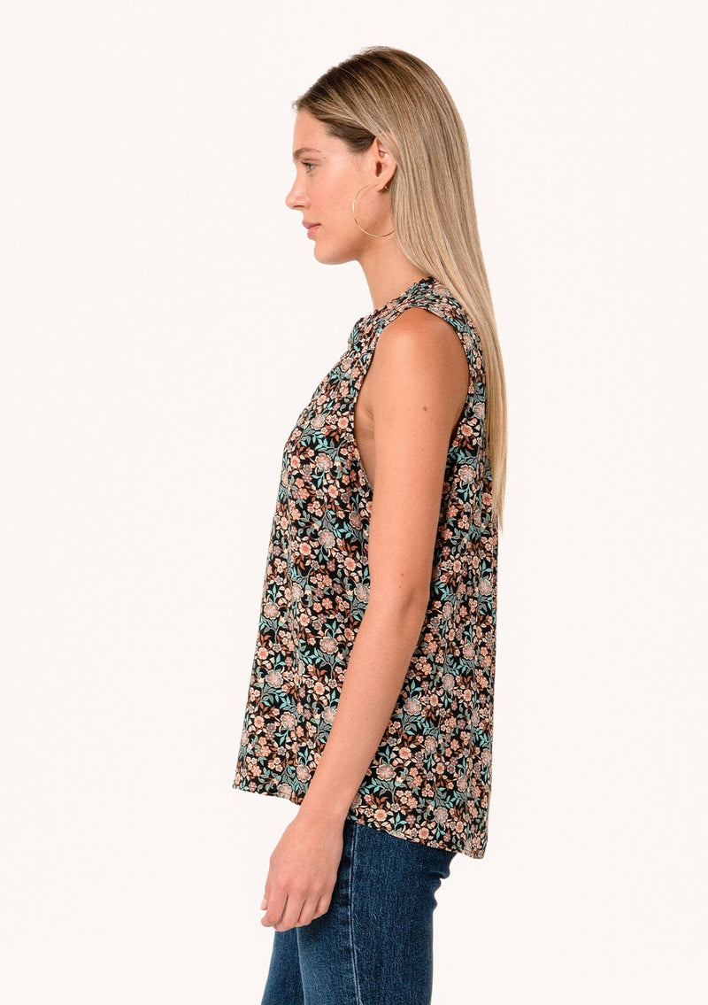 [Color: Black/Teal] A side facing image of a blonde model wearing a transitional fall sleeveless blouse in a black and teal floral print. With a button front, a ruffle trimmed neckline, and a relaxed fit. 