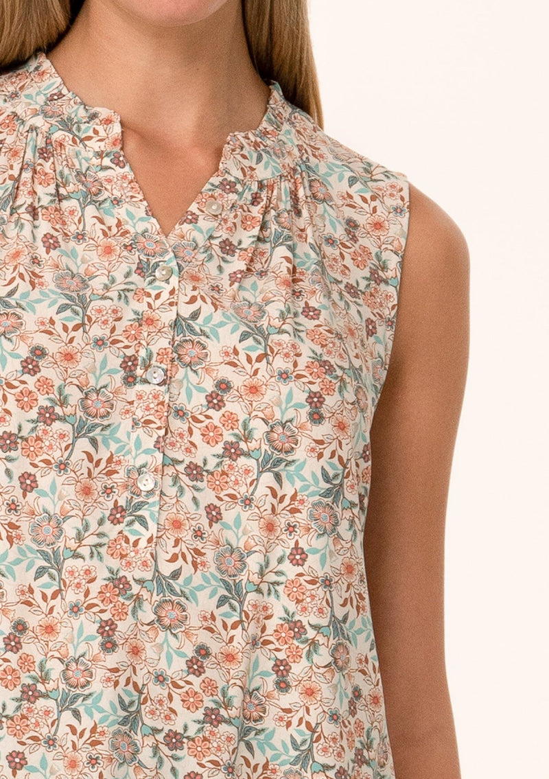 [Color: Cream/Teal] A close up front facing image of a blonde model wearing a transitional fall sleeveless blouse in a cream and teal floral print. With a button front, a ruffle trimmed neckline, and a relaxed fit. 