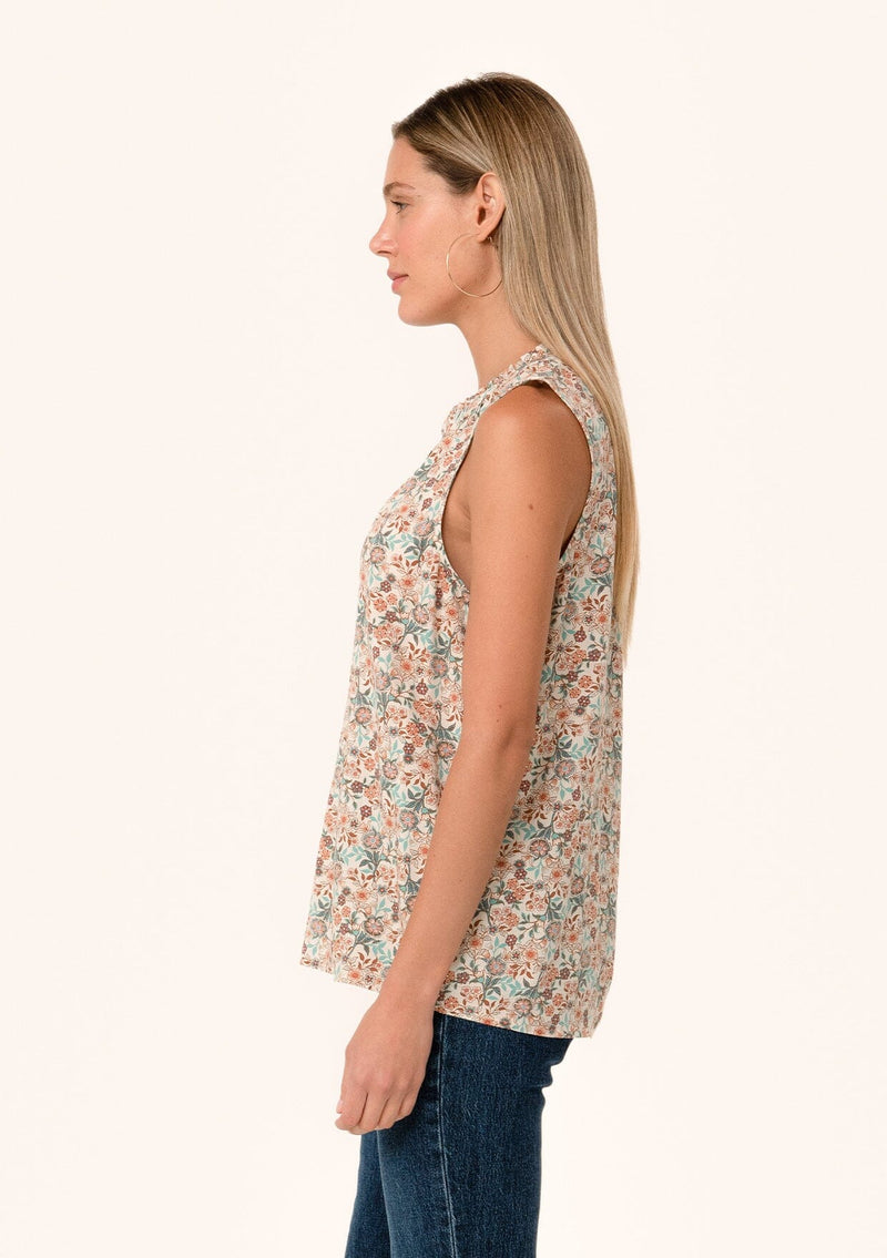 [Color: Cream/Teal] A side facing image of a blonde model wearing a transitional fall sleeveless blouse in a cream and teal floral print. With a button front, a ruffle trimmed neckline, and a relaxed fit. 